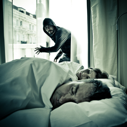 Encapuchado con barba y sudadera negra entra por la ventana de una casa con un hombre y una mujer dormidos en la cama.