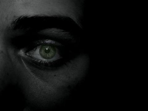 Cara de una persona en la oscuridad en la que solo se ve un ojo, de color verde, con expresión asustada.